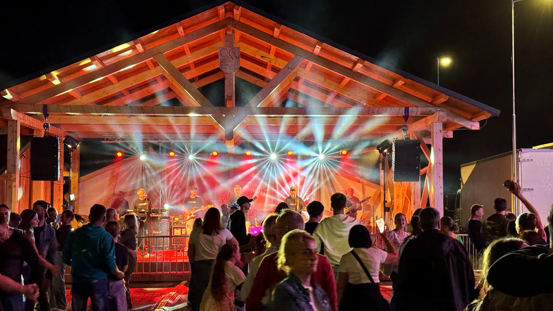 Kapela na drevenom amfiteátri s nasvietením a line array ozvučením, dav pred pódiom